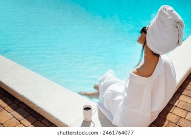 Young pretty woman in a white bathrobe by the swimming pool and holds a cup of coffee. Relaxed caucasian female in sunglasses and a towel on her head sits at the poolside. Vacation and spa concept. - Powered by Shutterstock