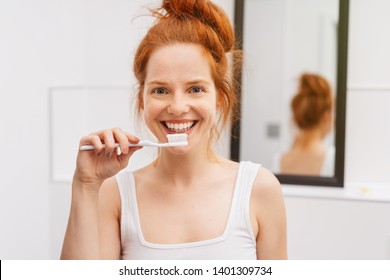 Young pretty woman in white shirt holding toothbrush before brushing her teeth looking at camera and smiling. Bust front portrait with reflection in the mirror blurred in background - Powered by Shutterstock