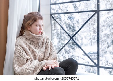 A Young Pretty Woman Without Gadgets Is Sitting By The Window In A Warm Beige Sweater And Admiring The Winter Forest.  Winter Vacation In A Country Hotel, Cozy Mood Concept
