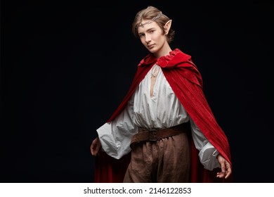 A Young Prince Of Elves In A Red Velvet Cloak And With A Crown On His Head. Studio Portrait On A Black Background With Copy Space. Fairy Tale, Magic. Fantasy.