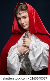 A Young Prince Of Elves In A Red Velvet Cloak And A Crown Stands With A Sword In His Hands. Studio Shot On A Black Background. Fairy Tale, Magic. Fantasy.