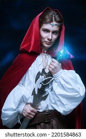 A Young Prince Of Elves In A Red Velvet Cloak And A Crown Stands With A Sword In His Hands. Starry Night Sky Background. Fairy Tale, Magic. Fantasy.