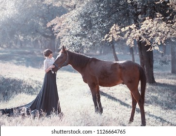A young princess in a vintage dress with a long train, with tenderness and love, hugs her horse. The brunette girl in a white blouse, black skirt. The background is fantastically beautiful forest