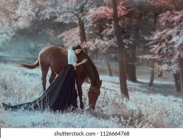 A young princess in a vintage dress with a long train, with tenderness and love, hugs her horse. The brunette girl in a white blouse, black skirt. The background is fantastically beautiful forest