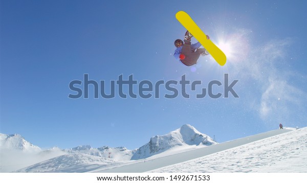 Young pro\
snowboarder riding the half pipe in big mountain snow park, jumping\
out of the halfpipe wall and over the sun, performing tricks and\
rotations with grabs in sunny\
winter