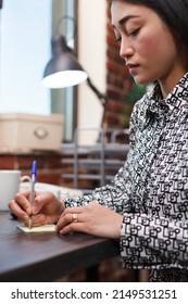 Young Productive Asian Businesswoman Taking Important Notes While Sitting At Desk In Company Workspace. Marketing Agency Employee Writing Down Client Personal Information Regarding Collaboration.