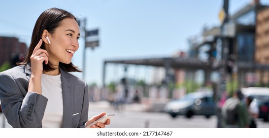 Young professional Asian business woman wearing suit using wireless earphones mobile internet data holding smartphone talking on mobile phone having chat on cellphone walking on city street. Banner