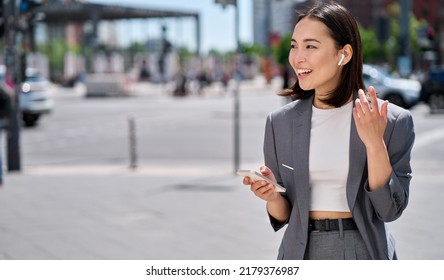 Young professional Asian business woman wearing suit using wireless earphones holding smartphone talking on mobile phone having chat on cellphone walking on urban city street. - Powered by Shutterstock