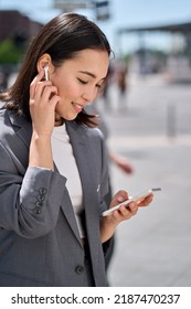 Young Professional Asian Business Woman Or Student Wearing Earphone Holding Smartphone Looking At Mobile Phone Having Video Chat On Cellphone, Watching Webinar Standing On City Street. Vertical