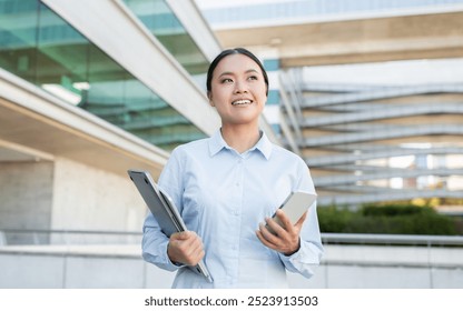 A young professional Asian lady is outdoors in a modern urban setting, smiling confidently while holding a tablet and smartphone. Buildings and greenery add context to her upbeat demeanor. - Powered by Shutterstock