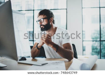 Young professional bearded designer wearing eye glasses working at modern loft studio-office with desktop computer.Blurred background. Horizontal