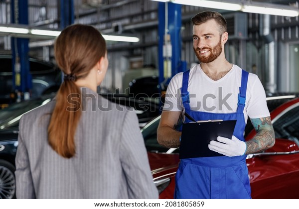 Young professional car mechanic man in denim\
blue overalls white t-shirt gloves hold clipboard with papers\
document talk with female manager work in light modern vehicle\
repair shop workshop\
indoors.
