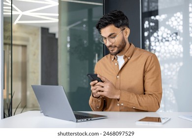 Young professional checking phone while working on laptop in modern office. Business setting, casual attire, modern technology. - Powered by Shutterstock