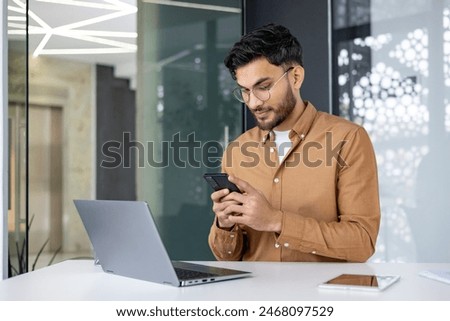 Young professional checking phone while working on laptop in modern office. Business setting, casual attire, modern technology.