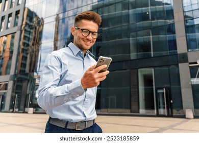 Young Professional Checking Smartphone While Standing Outside Modern Office Building in Downtown Area During Bright Sunny Day