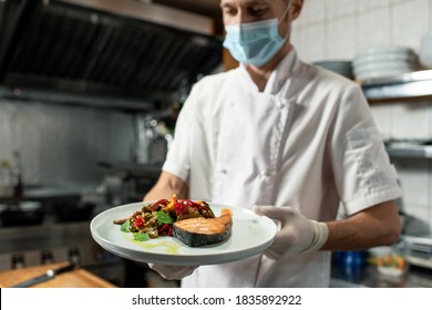 Young Professional Chef In White Uniform And Protective Mask And Gloves Holding Plate With Fried Salmon And Vegetable Garnish In The Kitchen