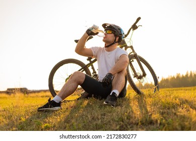 Young professional cyclist dressed in cycling clothing and protective helmet drinking water from the bottle. Active healthy lifestyle. Sports man resting after exercise in nature. Mountain bike.
