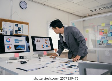 Young professional examines data visualized in colorful charts displayed on computer monitors and printed documents, optimizing business strategies and performance