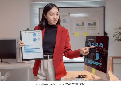 Young professional explaining data analysis during a business meeting, holding clipboard and pointing at computer screen showing charts and graphs