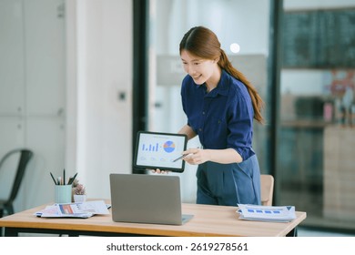 Young professional explaining data analysis during a business meeting, holding a tablet and pointing at a computer screen displaying charts and graphs.