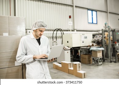 Young Professional Is Leaning On The Boxes In Warehouse For Food Packaging. Manager Is Working On The Computer In Automated Production Line At Modern Factory. Color Toned Image.