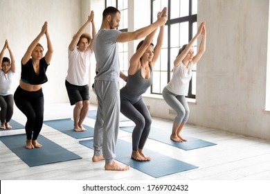 Young Professional Male Yoga Instructor Correcting Chair Position Performance Of Motivated Female Students At Group Morning Class Indoors, Fit Sporty People In Activewear Practicing Utkatasana Pose.