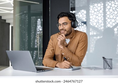 Young professional man wearing headphones and taking notes during a video conference in a modern office setting.