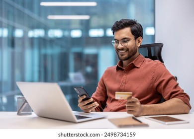 Young professional in a modern office using a phone and credit card for an online transaction. Displaying happiness and success in a business environment. - Powered by Shutterstock