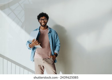 Young professional on a balcony in a modern office. He holds a laptop and smiles, embodying the success of a startup entrepreneur. Dressed in business casual attire,he exudes confidence and creativity - Powered by Shutterstock