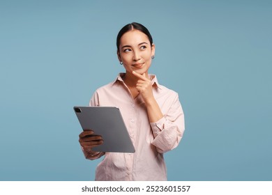 Young professional, smiling Asian women holding digital tablet, looking away, standing isolated on blue background. Advertisement, online technology concept