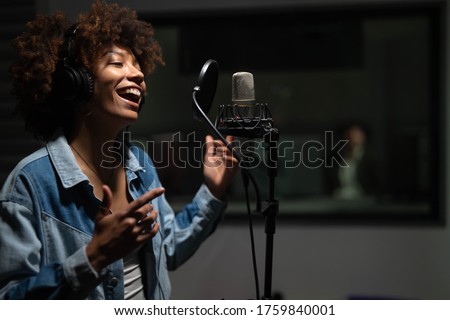 An young professional smiling energetic african female singer wearing headphones is performing a new song with a microphone while recording it in a music studio. 