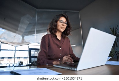 Young professional it specialist latin hispanic business lady working on laptop pc sitting at desk in modern office space. 30s middle eastern indian woman using computer technology app for work online