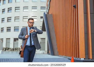 A young professional in a suit checks his watch while walking in an urban business district. The modern architecture and his focused expression suggest a busy and dynamic work environment. - Powered by Shutterstock