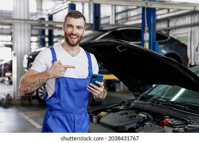 Young Professional Technician Car Mechanic Man In Blue Overalls White T-shirt Use Hold Point Finger On Mobile Cell Phone Fix Problem With Raised Hood Bonnet Work In Vehicle Repair Shop Workshop Indoor