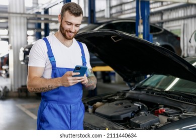 Young Professional Technician Car Mechanic Man 20s In Blue Overalls White T-shirt Use Hold Mobile Cell Phone Browsing Fix Problem With Raised Hood Bonnet Work In Vehicle Repair Shop Workshop Indoor.