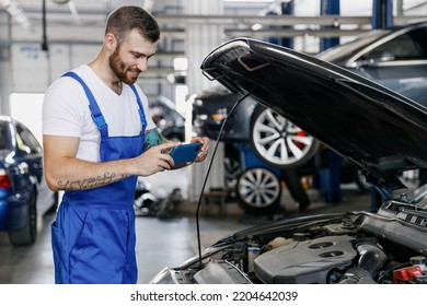Young professional technician car mechanic man in blue overalls white t-shirt take photo picture on mobile cell phone fix problem with raised hood bonnet work in vehicle repair shop workshop indoor.