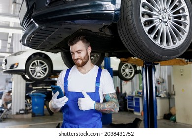 Young Professional Technician Mechanic Man In Blue Overalls T-shirt Use Hold Mobile Cell Phone Show Thumb Up Stand Near Car Lift Check Technical Condition Work In Vehicle Repair Shop Workshop Indoors