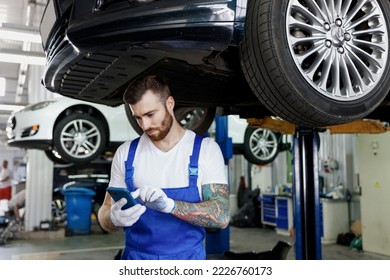 Young professional technician mechanic man wearing denim blue overalls t-shirt use hold mobile cell phone stand near car lift check technical condition work in vehicle repair shop workshop indoors. - Shutterstock ID 2226760173