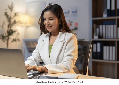 Young professional woman sitting at a desk, typing on her laptop while smiling and working late into the night, surrounded by warm office lighting that enhances her focused ambiance - Powered by Shutterstock