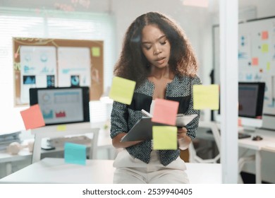 Young professional woman working with sticky notes and clipboard, analyzing charts and data in a bright modern office - Powered by Shutterstock