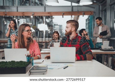 Young professionals collaborate at a modern open-plan office desk, using laptops and notebooks while chatting and sharing ideas. Casual startup atmosphere, teamwork, creativity and productive coworking scene.