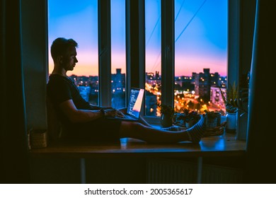 A young programmer is coding on a laptop in the dark sitting on a window with a view of the lights of the night city, colored lighting in the room, home decor - Powered by Shutterstock - Get 15% off with code: PIKWIZARD15