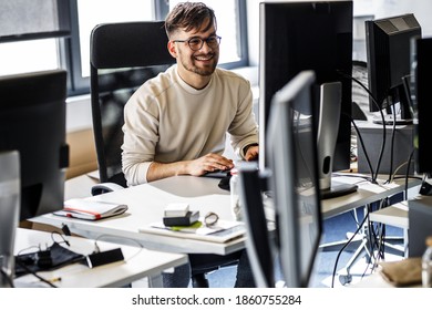 Young Programmer Sitting At The Desk In His Office And Working .He Talk With Someone And Smiling.	
