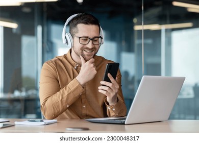 Young programmer working inside office with laptop, man in headphones smiling and listening to online music, audio podcast and audiobook, businessman in shirt at workplace holding phone. - Powered by Shutterstock