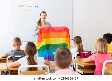 Young progressive female teacher discussing with preteen children about LGBT social movements in classroom, holding rainbow flag