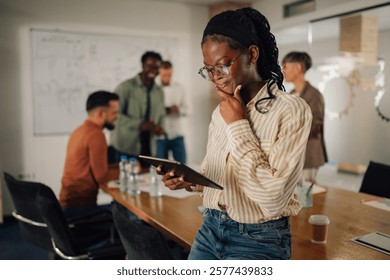 Young project manager is using digital tablet and thinking about new business ideas during a meeting with her diverse team in the conference room