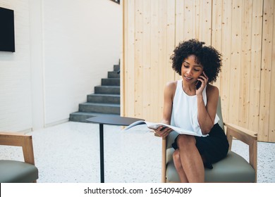 Young prosperous dark skinned woman having mobile phone conversations about business while checking finance news from magazine, smiling african american female editor in elegant wear making call