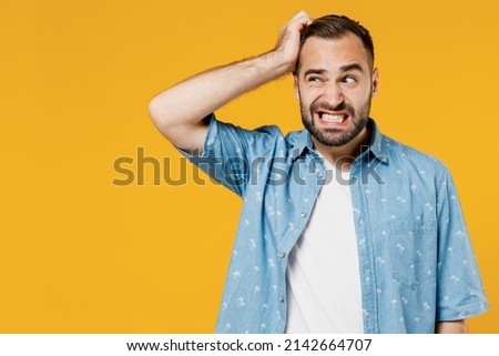 Young puzzled embarrassed bewildered caucasian man 20s wearing blue shirt white t-shirt scratch head look aside say oops isolated on plain yellow background studio portrait. People lifestyle concept