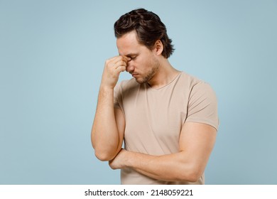 Young puzzled pensive thoughtful sad troubled man in casual basic beige t-shirt pinching rub put hand on nose bridge isolated on pastel blue color background studio portrait. People lifestyle concept