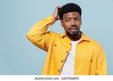 Young puzzled sad man of African American ethnicity wear yellow shirt look aside on workspace scratch hold head isolated on plain pastel light blue background studio portrait. People lifestyle concept